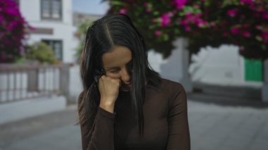 Arka planda çiçekleri olan İspanyol bir kadın, doğal bir ortamda yorgunluğunu ya da tefekkürünü ifade ediyor, koyu renk bir kıyafet giyiyor..