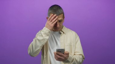 Akıllı telefonlu bir adam, izole edilmiş mor duvara şaşırmış görünüyor. Ağzı derin derin dokunuyor..