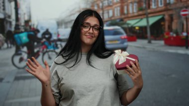 Yoğun şehir caddesinde elinde kalp şeklinde hediye kutusu olan bir kadın. Arka planda şehir yaşam tarzını gösteren bisikletçiler var..