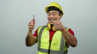İnşaat malzemeleri ve beyaz arka planda sert bir şapka takan genç adam kendine güvenen bir gülümseme ve onaylanmış bir jest içeren bir cihaz sergiliyor.