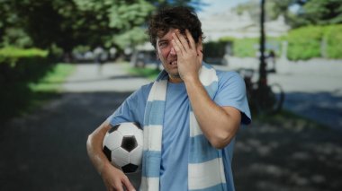 Parkta futbol topu tutan İspanyol bir adam sinirli görünüyor, mavi gömlek ve atkı giyiyor, yeşil ağaçlarla çevrili ve arka planda bulanıklık var, açık hava sporları yaptığını gösteriyor..