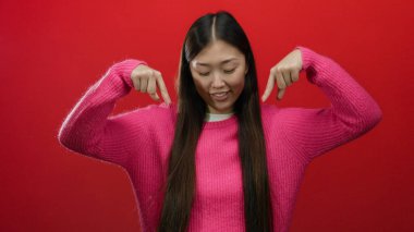 Pembe süveter giyen, uzun siyah saçlı, koyu kırmızı arka planda gülümseyen, aşağıya bakan, neşeli bir ifade ve canlı bir jest tasvir eden bir kadın..