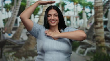 Tatil köyündeki kadın, canlı bir atmosferi olan palmiye ağaçlarının altında neşeli bir şekilde poz verirken el hareketleri yapıyor..