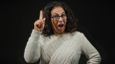 Kıvırcık saçlı gözlüklü İspanyol kadın şaşkınlığını dile getiriyor ve siyah arka planda bir fikir belirterek parmağını kaldırdı..