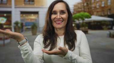 Şehir sokaklarında gülümseyen İspanyol bir kadın iki eliyle şehir geçmişine doğru el kol hareketi yapıyor..
