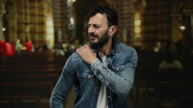 Kot ceketli, sakallı bir İspanyol Katolik kilisesinde duruyor ve omzunu tutarken rahatsız olduğunu söylüyor..