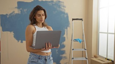 Modern oturma odasında merdivenle dizüstü bilgisayar kullanan ve duvarları boyayan bir kadın, yeni bir apartman dairesinde sıradan kıyafetler giymiş genç ve çekici bir esmer kadını sergiliyor..