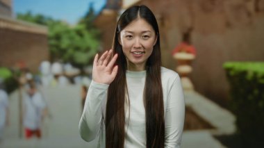 Gülümseyen ve el sallayan bir kadın ağaçlarla ve arka plandaki insanlarla dolu parlak bir şehir gününü gözler önüne seriyor..