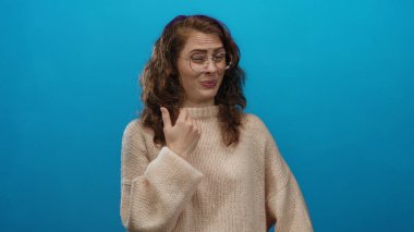 İzole edilmiş mavi arka planda, rahat kazak giyen kıvırcık saçlı ve gözlüklü bir kadın tiksindirici bir yüz ifadesi sergiliyor.