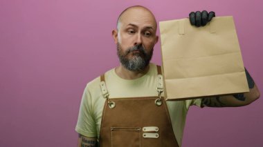 Önlüklü kel sakallı adam pembe arka planda ifadesiz yüzü olan kağıt torba tutuyor..