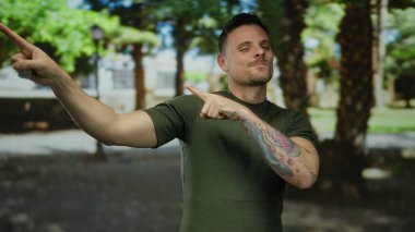 Dövmeleri olan genç bir adam gülümsüyor ve yemyeşil bir parkı işaret ediyor. Açık havada gündelik bir anı yakalıyor..