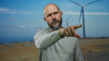 Sakallı ve kel kafalı bir adam tarlada duruyor. Arka planda rüzgar türbinleriyle gündüz vakti bulutlu gökyüzünün altında.