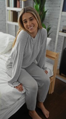 Kadın rahat bir yatak odasında oturuyor, gülümsüyor, gevşiyor, sarı saçlar, günlük kıyafetler, huzurlu ev ortamları, ev ortamları, sakin atmosfer..