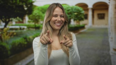 Sarışın kadın, kampüs manastırında beyaz bluz giyen kamerayı işaret ediyor. Oyuncu özgüveni, nişanlı arkadaşlığı. Eğlenceli mutluluk bağlantısı, spontanlık sıcaklığını davet ediyor..