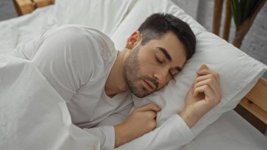 Genç bir adam rahat bir yatak odasında beyaz çarşaflarla huzurlu bir şekilde uyuyor. Sakin bir ev ortamında huzur ve huzuru ön plana çıkarıyor..