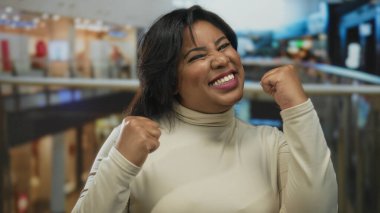 Canlı bir alışveriş merkezinde parmakları havada gülümseyen, kendine güveni ve neşe saçan, krem bir bluz giyen ve kapalı bir yerde, kalabalık dükkanların ortasında duran bir kadın..