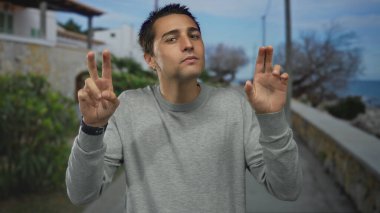 Gri kazaklı adam deniz kenarında iki eliyle tırnak işareti yapıyor; şüphecilik.