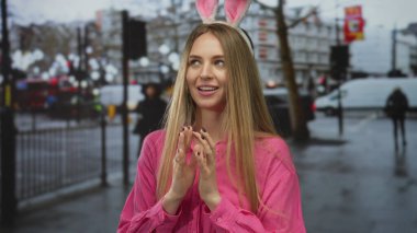 Şehir arka planında pembe giyinmiş bir sokakta tavşan kulağı takan bir kadın hareketli bir ortamda neşeli paskalya titreşimleri yayıyor..