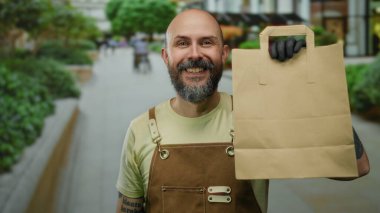 Kentsel caddede sakallı bir adam, tulum ve eldiven giyip, kağıt torba tutuyor..