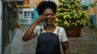 Önlükle gülümseyen bir kadın arka planda şenlik ışıkları olan dekore edilmiş bir şehir caddesinde barış işareti yapıyor..