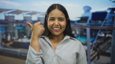 Genç, Latin kökenli, güvertede, bulanık deniz arka planına gülümseyen bir kadın başparmağı..