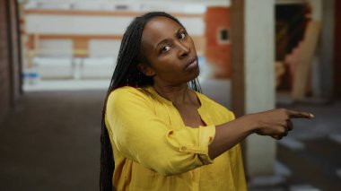 Bitmemiş bir endüstriyel binada, üzerinde ciddi bir ifade olan sarı bir tişört olan bir inşaat alanını işaret eden bir kadın.