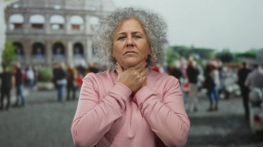 Pembe kazaklı, gri saçlı bir kadın İtalya 'da Roma Kolezyumu' nun önünde boğazını tutuyor..