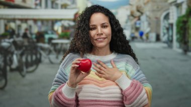 Dışarıda gülümseyen İspanyol kadın renkli bir kazak içinde kırmızı bir kalple kalabalık bir caddede neşe ve sıcaklık öneriyor..