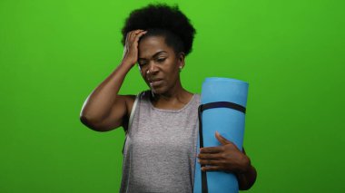 Mavi yoga minderi tutan kadın, yeşil arka plana karşı gergin, afro saç stili, kolsuz bluz giyiyor, sakin ve düşünceli..