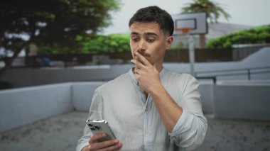 Akıllı telefonlu genç adam bir basketbol sahasında dikiliyor, derin derin düşünüyor, ağaçlar ve arka planda bir çemberle..