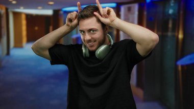 Sakallı, kulaklık takan, otel koridorunda boynuz hareketi yapan, neşeli ve kapalı mekan sahnesini anlatan genç bir adam..