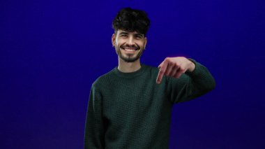 Genç adam kendinden emin bir şekilde gülümserken izole edilmiş mavi bir arkaplanı işaret ederek dostça ve davetkar bir tavır sergiliyor..