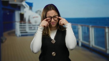 Güneşli bir günde arka planda okyanus olan bir yolcu gemisinde, rahat bir ifadeyle kameraya gülümseyen, günlük kıyafetler ve gözlük takan bir kadın..