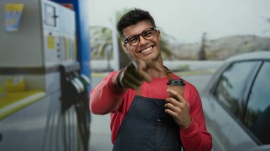 Kırmızı kazaklı, gülümseyen İspanyol bir adam benzin istasyonunda kahve tutuyor, arka planda modern araba ve palmiye ağaçları var..