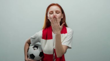 Futbol topu tutan kızıl saçlı bir kadın kırmızı ve beyaz bir eşarp takarak öpüşüyor, düz beyaz bir arkaplanda duruyor..
