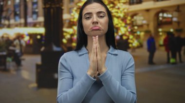 Üzgün ifadeli ve elleri olan genç bir kadın, arka planda şenlik ışıkları olan kalabalık bir caddede gece yalvaran bir jestte sıkıca tutundu..