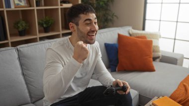 Ev ortamında video oyunları oynayan genç adam zaferi gülümseyerek kutluyor. Etrafı rahat bir dekor ve rahat bir atmosferle çevrili..