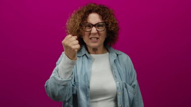 Orta yaşlı bir kadın, canlı pembe arka plana karşı öfkesini dışa vuruyor, gözlük ve kot gömlek giyiyor, hayal kırıklığı ve kararlılık duygularını iletiyor.