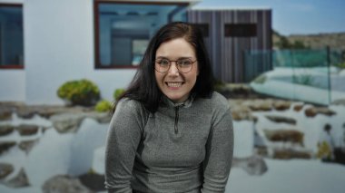 Modern lüks bir evin önünde, sakin bir gülümseme, esmer saçlar ve gözlüklerle, zarafet ve rahatlığı somutlaştıran, kendinden emin bir şekilde duran bir kadın..