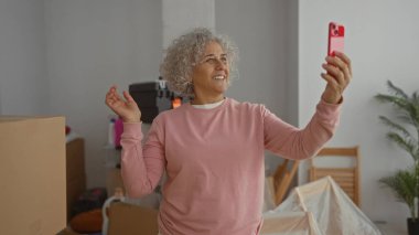 Pembe kazaklı kadın, yeni oturma odasındaki telefon görüşmesinde gülümsüyor. Etrafı kutular ve bitkilerle çevrili. Akıllı telefondan neşeli anlar yakalıyor..