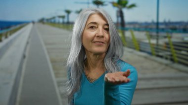 Gri saçlı yaşlı kadın deniz kenarında bir öpücük yolluyor, sakin bir kumsal sahili anını yakalıyor açık gökyüzünün altında rahat ve neşeli bir atmosferde..