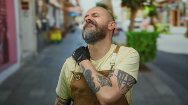 Şehir caddesinde sakallı bir adam, önlük ve eldiven giyiyor, güneşli bir günde, şehirde yorgunluk gösteriyor..