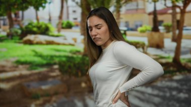Sırtı ağrıyan genç bir kadın açık hava parkı ortamında duruyor, canlı yeşillikler ve ağaçların arasında huzursuzluğunu dile getiriyor, doğal gün ışığı altında uzun kollu beyaz bir bluz giyiyor..