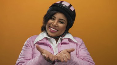 Kask takan ve pembe ceketli bir kadın sıcak bir şekilde gülümsüyor. Arkaplan olarak turuncu bir duvarla kameraya doğru uzanan elleri uzatıyordu..