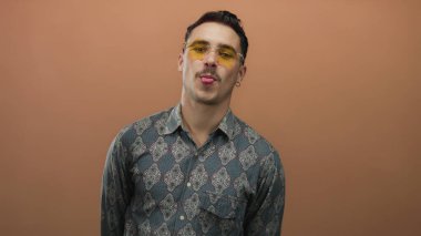 Güneş gözlüklü, desenli tişörtlü genç İspanyol bir adam kahverengi duvarın arkasına komik bir şekilde dil uzatıyor, izole bir çerçevede, tavır ve stil ifade ediyor..