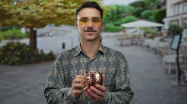 Genç adam gülümsüyor ve canlı bir şehir caddesinde fincanı tutuyor, şık gözlükler takıyor ve gür ağaçların altında desenli gömlek giyiyor, rahat bir şehir havası yayıyor..