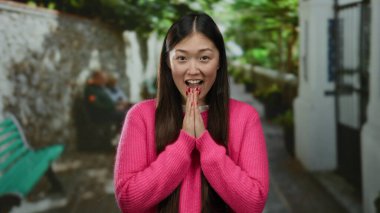 Kentsel sokak ortamında gülümseyen bir kadın pembe giyinmiş, yeşillikle mutluluğu ifade eden ve parlak bir günde bulanık arka planda oturan insanlar..