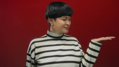 Canlı kırmızı arka planda yalnız gülümseyen bir kadın çizgili süveter giyiyor. Neşeli bir ifade sergiliyor. El hareketleriyle sunum ya da davetiye öneriyor..