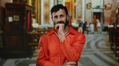 Turuncu tulumlu İspanyol sakallı bir adam Katolik kilisesinin içinde duruyor, içsel olarak poz veriyor, inanç, kefaret ve iç gözlem temaları öneriyor..
