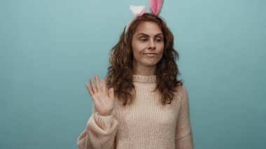 Pembe kazak giyen ve tavşan kulaklı bir kadın mavi arka planda şakacı bir şekilde el sallayarak eğlenceli ve misafirperver bir ortam yaratıyor..